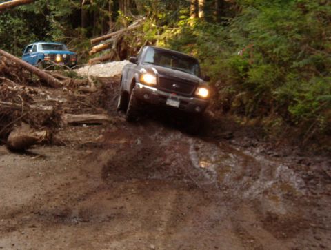 Dark pickup truck driving down a muddy off-road trail with a blue SUV behind it.