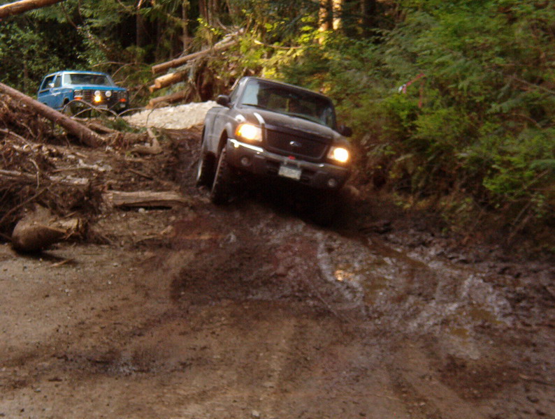 Dark pickup truck driving down a muddy off-road trail with a blue SUV behind it.