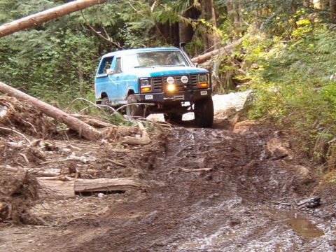 Blue 1985 Ford Bronco off-roading on a muddy, wooded trail.