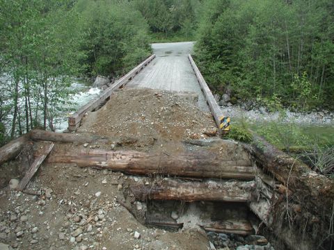 Damaged wooden bridge over a river with exposed logs and dirt.