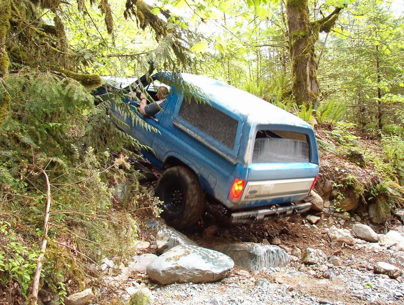 Blue Ford Bronco climbing a steep, rocky, wooded trail.