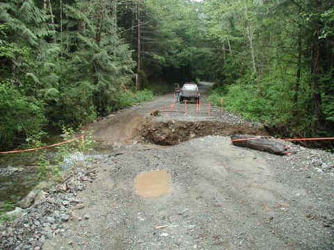 Silver SUV on a damaged forest road with orange safety tape and a washed-out section.