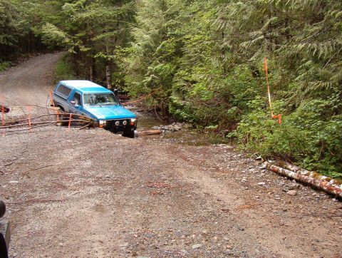 Blue Ford Bronco stuck in a stream on a gravel road with orange flagging.