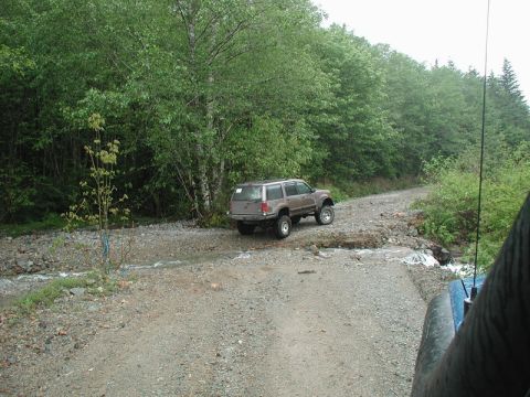 Lifted tan SUV crossing a shallow stream on a gravel road.