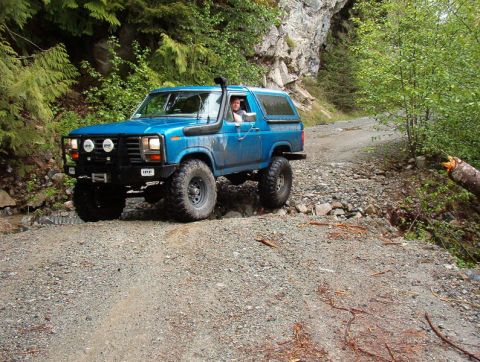 Blue lifted 1980s Ford Bronco driving on a rocky off-road trail.