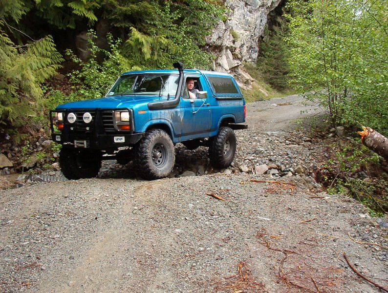 Blue lifted 1980s Ford Bronco driving on a rocky off-road trail.
