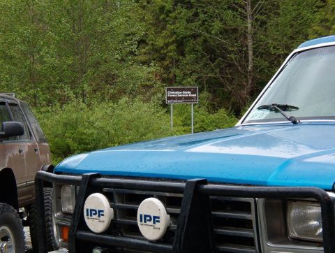 Blue 1985 Ford Bronco with bull bar and IPF lights, parked next to a brown SUV near a forest service road sign