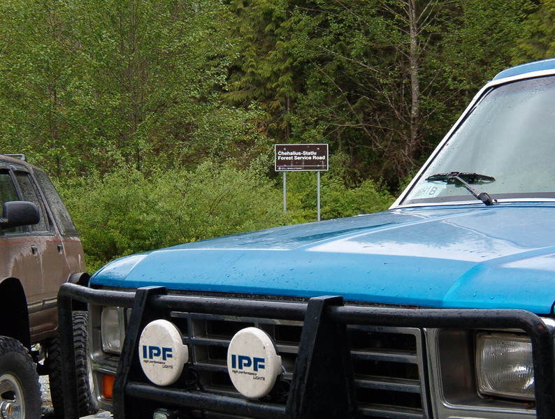 Blue 1985 Ford Bronco with bull bar and IPF lights, parked next to a brown SUV near a forest service road sign.