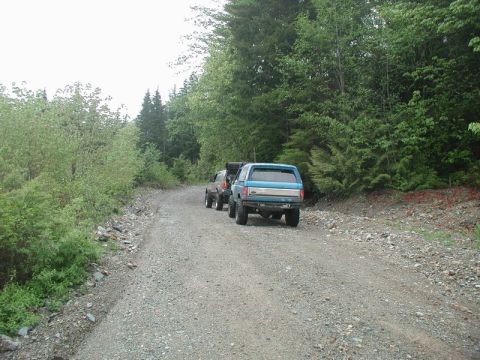 Blue Ford Bronco with camper shell on a gravel road next to trees.