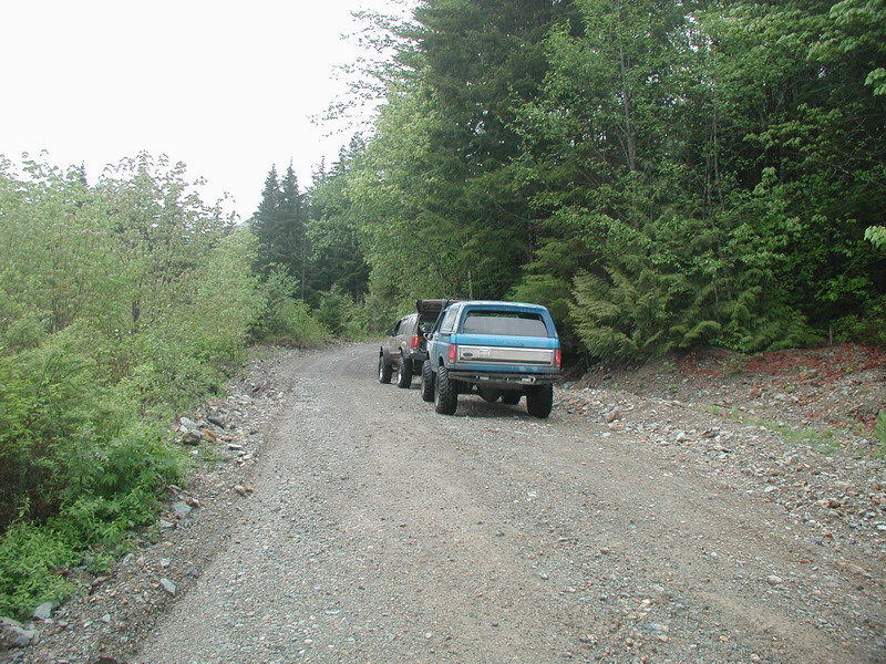 Blue Ford Bronco with camper shell on a gravel road next to trees.