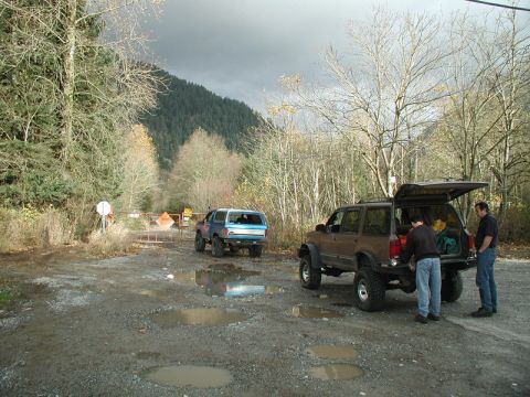 Two modified SUVs parked on a gravel road with a gate and construction signs ahead. Men loading gear into one
