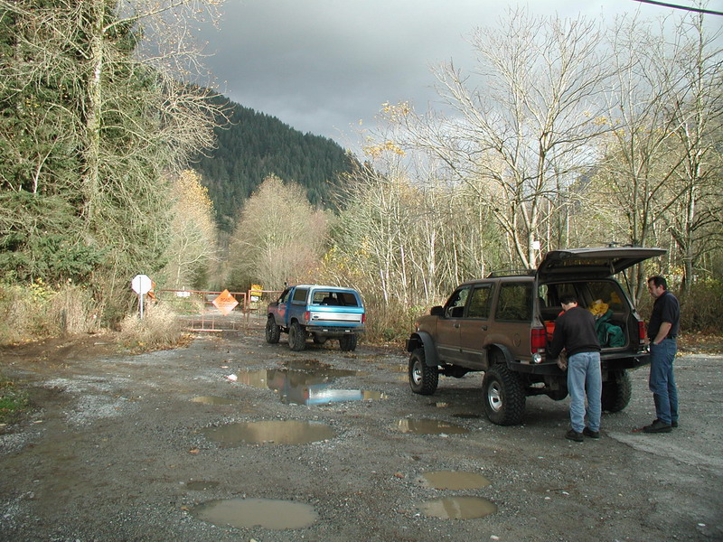 Two modified SUVs parked on a gravel road with a gate and construction signs ahead. Men loading gear into one SUV.