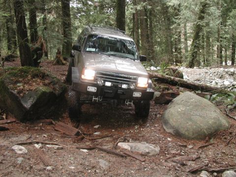 Tan SUV off-roading on a forest trail, going over a log and a boulder.