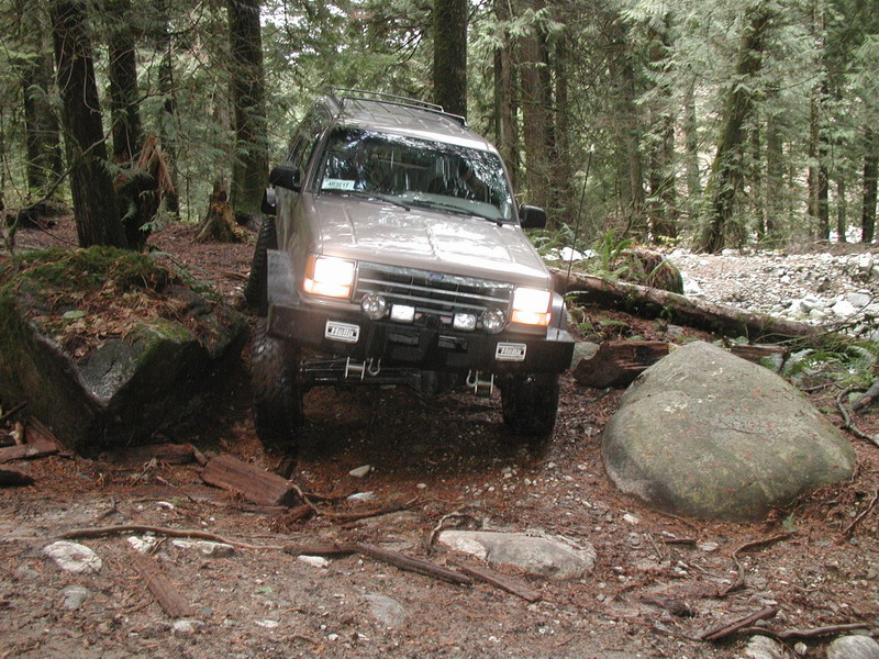 Tan SUV off-roading on a forest trail, going over a log and a boulder.
