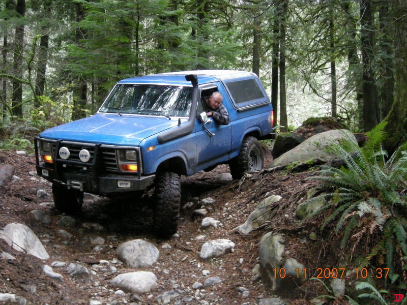 Blue 1985 Ford Bronco off-roading on a rocky trail in a forest.