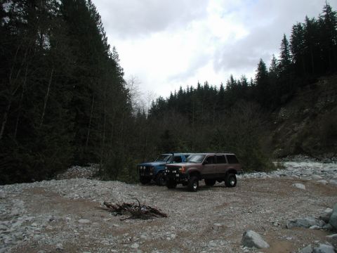 Two SUVs parked in a forest clearing by a river, with firewood in the foreground.