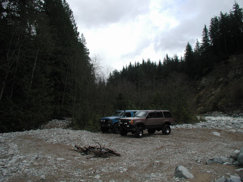 Two SUVs parked in a forest clearing by a river, with firewood in the foreground.