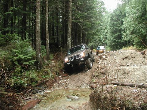 Two off-road SUVs on a muddy forest trail, one climbing a hill over a stream.