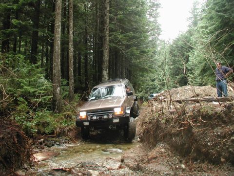 Tan Ford Bronco driving through a stream in a forest.