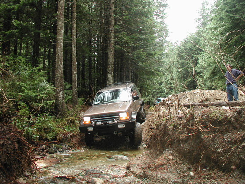 Tan Ford Bronco driving through a stream in a forest.
