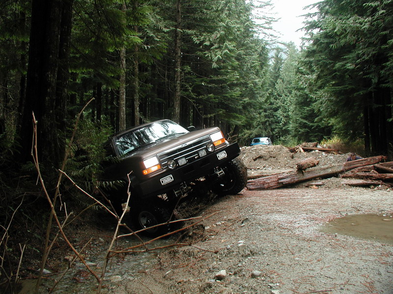 Brown Ford Bronco on a muddy forest trail with logs and another SUV in the distance.
