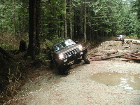 Brown 1985 Ford Bronco off-roading on a muddy trail in a forest.
