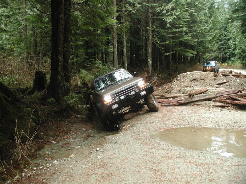 Brown 1985 Ford Bronco off-roading on a muddy trail in a forest.