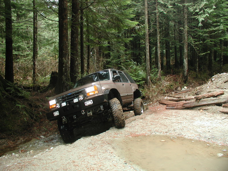 Lifted tan Ford Explorer off-roading in a forest, climbing out of a muddy rut.