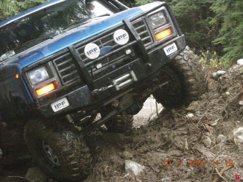 Blue off-road vehicle climbing a muddy hill with trees in the background.