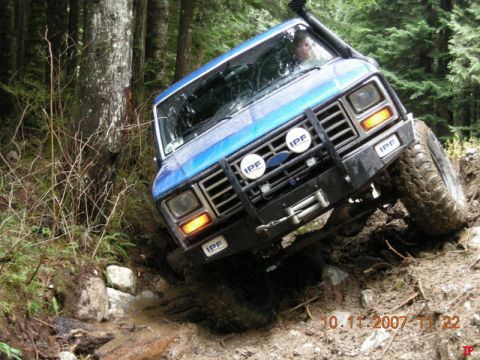Blue 1985 Ford Bronco off-roading on a muddy trail in a forest.