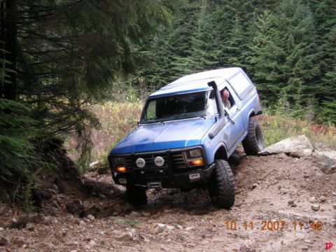 Blue 1985 Ford Bronco off-roading on a rocky trail in a forest.