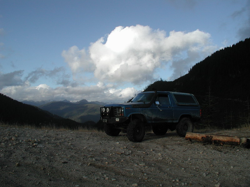 Blue Ford Bronco parked on a gravel overlook with mountains and clouds.