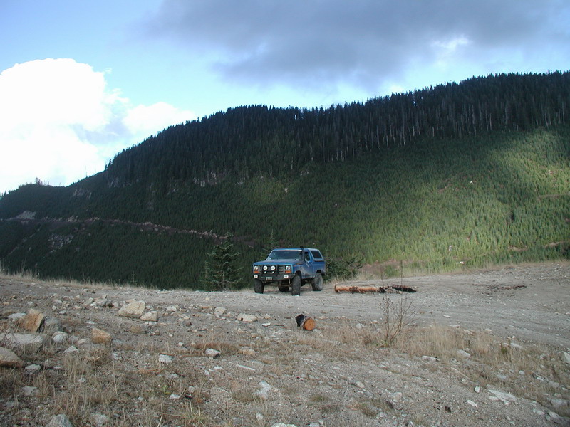 Blue 1985 Ford Bronco parked on a mountain clearing with a forest backdrop.