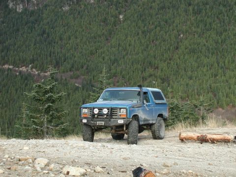 Blue 1980s Ford Bronco with off-road modifications parked on gravel, forest mountain background.