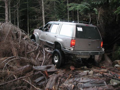 Lifted SUV driving over fallen trees and debris in a forest.