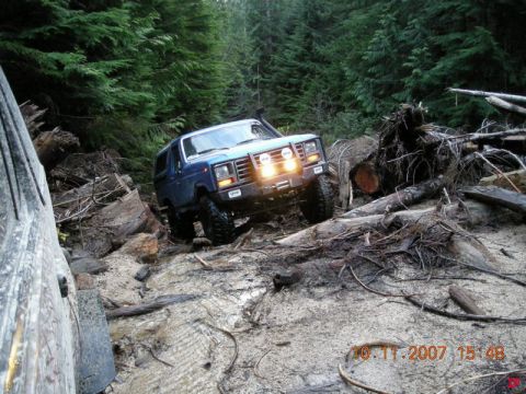 Blue 1985 Ford Bronco off-roading through a forest with fallen trees and mud.