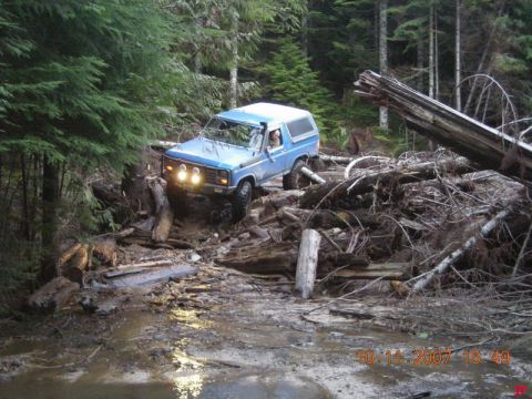 Blue 1980s Ford Bronco off-roading through a muddy forest trail with fallen trees.