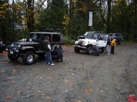 Two Jeep Wranglers parked in a wooded area with people gathered around one of the vehicles.