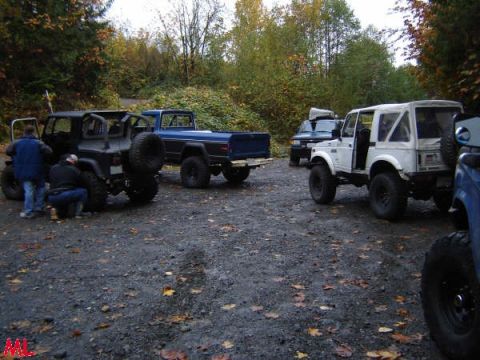 Off-road vehicles parked on gravel lot in autumn, including a blue Ford pickup and black Jeep.
