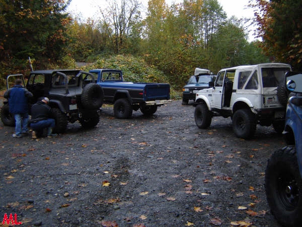 Off-road vehicles parked on gravel lot in autumn, including a blue Ford pickup and black Jeep.