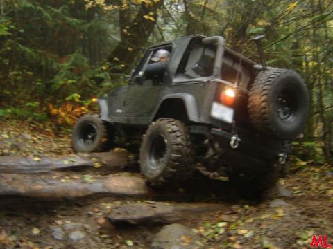 Jeep Wrangler off-roading over logs and rocks in a forest.