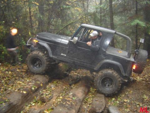 Black Jeep Wrangler off-roading over logs on a muddy trail in a forest.