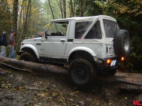 White soft-top Jeep Wrangler driving over a log on a muddy trail.