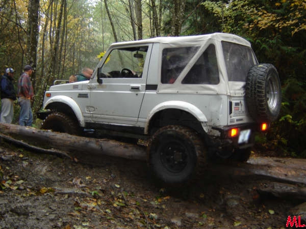 White soft-top Jeep Wrangler driving over a log on a muddy trail.