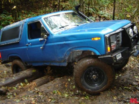 Blue 1985 Ford Bronco off-roading over logs and mud.