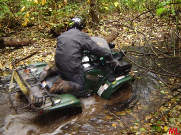 Person on an olive green ATV riding through a muddy stream in a forest.