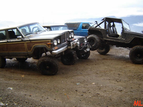 Lifted Jeep Wrangler and wood-paneled station wagon parked on dirt.