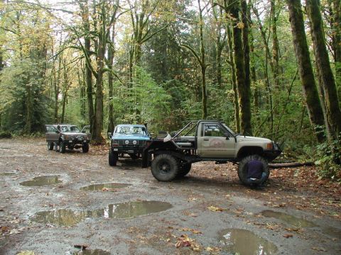 Three off-road trucks parked in a muddy forest clearing with a person working on a tire.