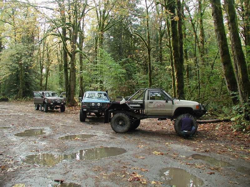 Three off-road trucks parked in a muddy forest clearing with a person working on a tire.