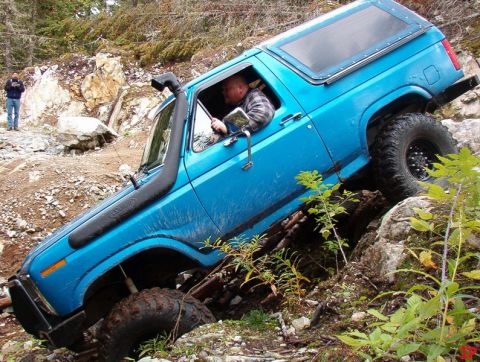Blue 1985 Ford Bronco off-roading on a rocky trail, tilted on its side.